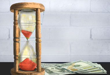 stylized hourglass with red sand sitting on top of a small stack of hundred dollar bills on a tabletop in a room with dull white walls